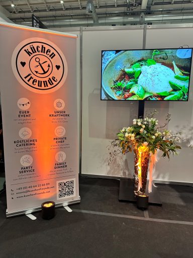 Stand mit Werbung, Bildschirm mit Speisen und Pflanze in einem modernen Messeumfeld.
